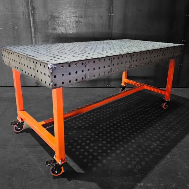 K&ouml;nnte beinhalten: Ein Metall-Arbeitstisch mit perforierter Oberseite und leuchtend orangefarbenen Beinen. Der Tisch ist rechteckig und steht auf R&auml;dern. Die Metalloberseite hat viele kleine L&ouml;cher. Die Beine sind leuchtend orange.