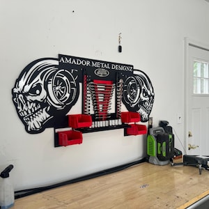 Puede incluir: Un organizador de herramientas de metal negro montado en la pared con dos diseños en forma de calavera y el texto "AMADOR METAL DESIGNZ". El organizador tiene contenedores de almacenamiento de plástico rojo y un estante para guardar herramientas.