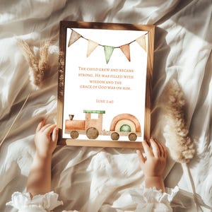 May include: A framed print featuring a watercolor illustration of a wooden train with a rainbow car. The print includes the text "THE CHILD GREW AND BECAME STRONG. HE WAS FILLED WITH WISDOM AND THE GRACE OF GOD WAS ON HIM. LUKE 2:40". The frame is held by a child.