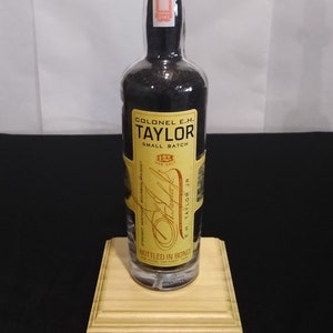 May include: A clear glass bottle lamp with a gold metal lampshade. The bottle is filled with black liquid and has a label that reads "Colonel E.H. Taylor Small Batch Bottled in Bond E.H. Taylor Jr."