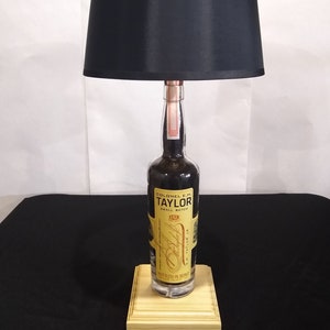 May include: A black lampshade with a wooden base. The base is made from a repurposed bottle of Colonel E.H. Taylor Jr. Small Batch Bourbon. The bottle label is yellow with black text.