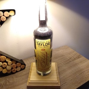 May include: A repurposed bottle lamp made from a Colonel E.H. Taylor Small Batch bourbon bottle. The bottle is filled with clear, sparkly stones and has a black lampshade. The bottle is sitting on a wooden base.
