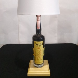 May include: A white lampshade sits atop a clear glass bottle with a black label that reads "Colonel E.H. Taylor Small Batch" and "Bottled in Bond". The bottle is on a wooden base.