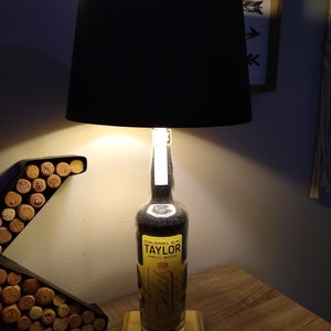 May include: A black lampshade with a clear glass bottle base filled with black sand. The bottle has a yellow label that reads "Colonel E.H. Taylor Small Batch" and "1887". The lamp is sitting on a wooden base.