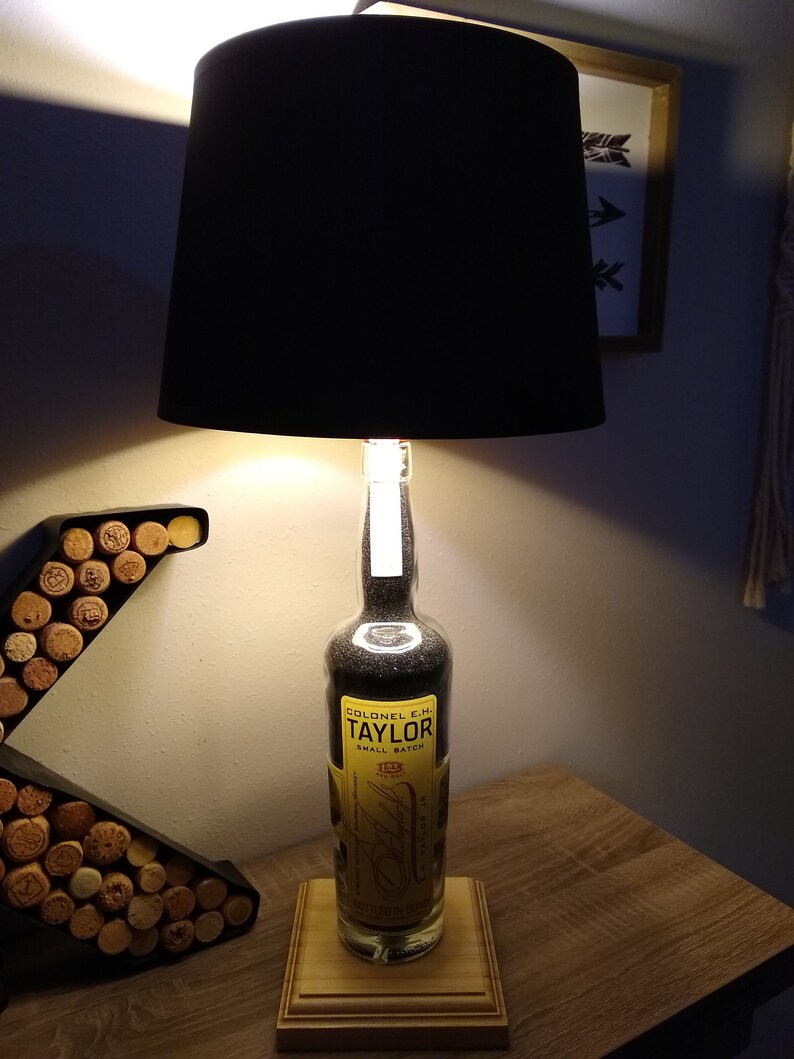 May include: A black lampshade with a clear glass bottle base filled with black sand. The bottle has a yellow label that reads "Colonel E.H. Taylor Small Batch" and "1887". The lamp is sitting on a wooden base.