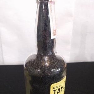 May include: A clear glass bottle filled with black sand. The bottle has a yellow label with the text "Colonel Taylor Small Batch Whiskey" printed on it.