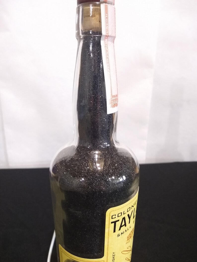 May include: A clear glass bottle filled with black sand. The bottle has a yellow label with the text "Colonel Taylor Small Batch Whiskey" printed on it.