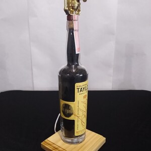 May include: A repurposed glass bottle lamp with a brass lampshade and a wooden base. The bottle is filled with black sand and has a label that reads "Colonel E. Taylor Jr. Small Batch Bourbon Whiskey Bottled in Bond."
