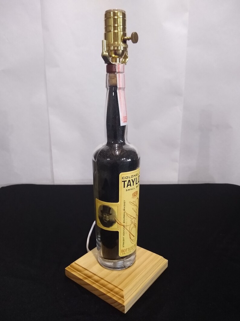 May include: A repurposed glass bottle lamp with a brass lampshade and a wooden base. The bottle is filled with black sand and has a label that reads "Colonel E. Taylor Jr. Small Batch Bourbon Whiskey Bottled in Bond."