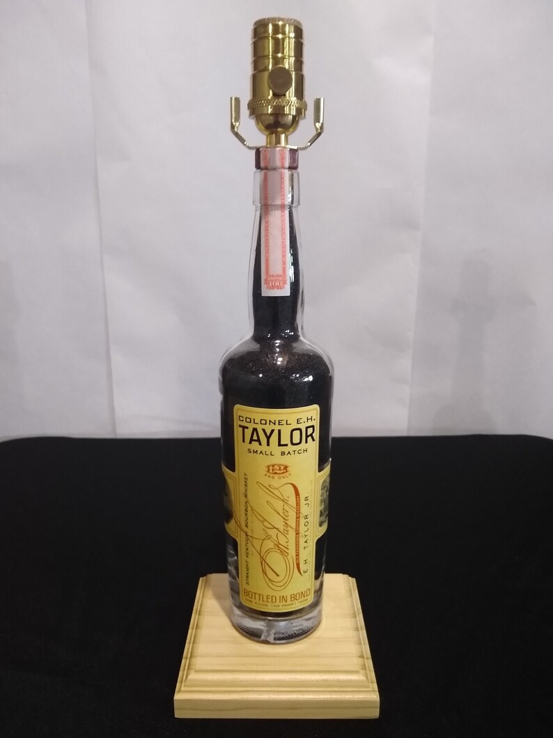 May include: A clear glass bottle lamp with a gold metal lampshade. The bottle is filled with black liquid and has a label that reads "Colonel E.H. Taylor Small Batch Bottled in Bond E.H. Taylor Jr."