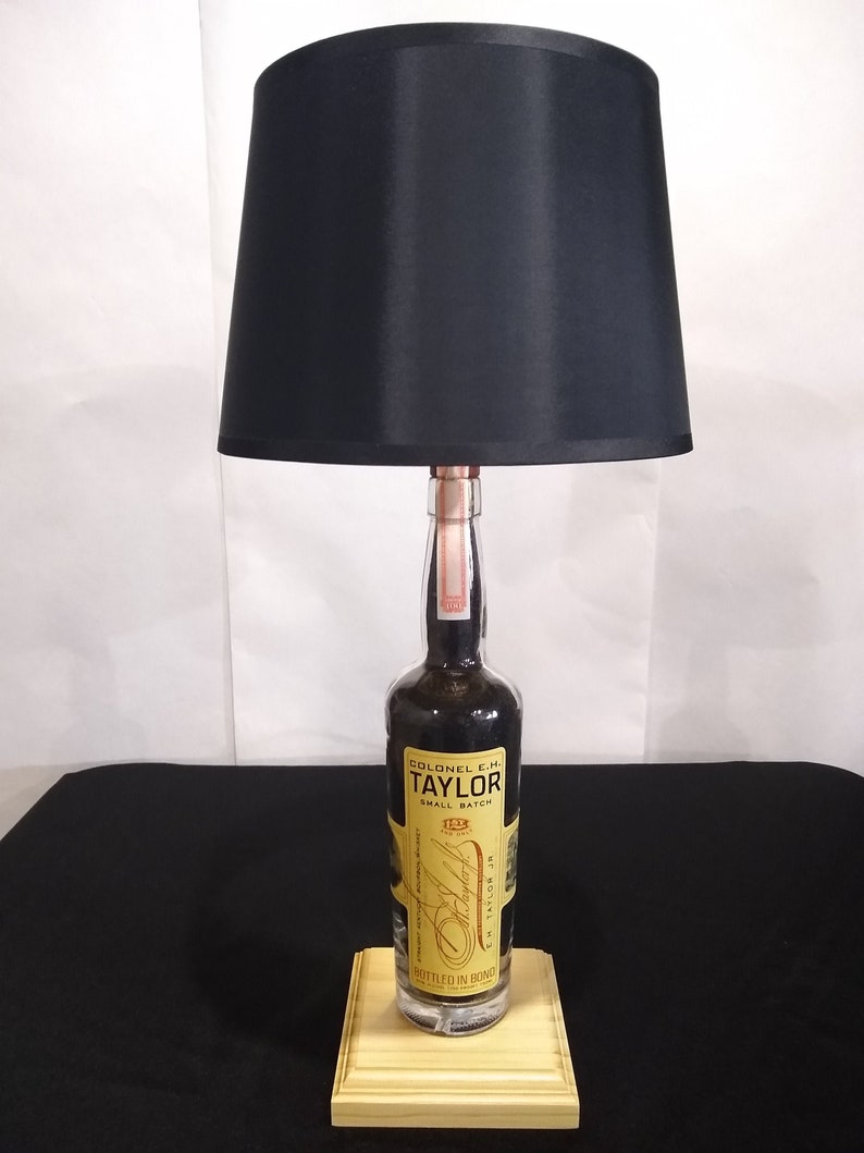 May include: A black lampshade with a wooden base. The base is made from a repurposed bottle of Colonel E.H. Taylor Jr. Small Batch Bourbon. The bottle label is yellow with black text.