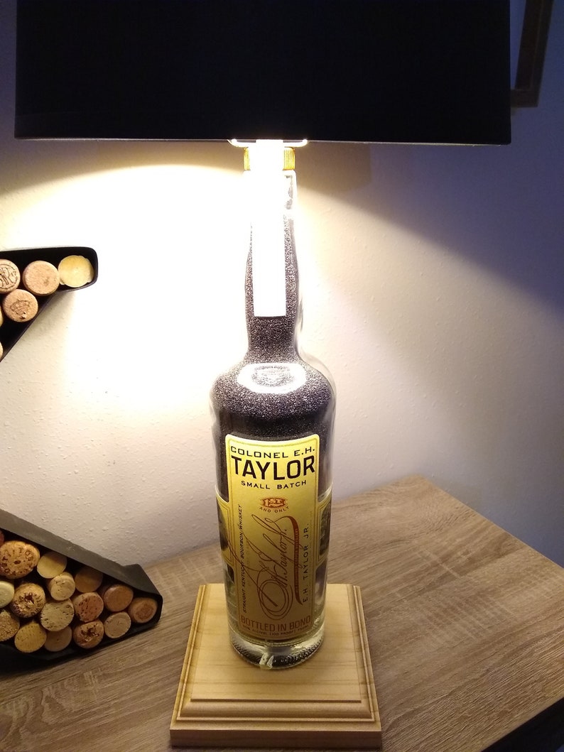 May include: A repurposed bottle lamp made from a Colonel E.H. Taylor Small Batch bourbon bottle. The bottle is filled with clear, sparkly stones and has a black lampshade. The bottle is sitting on a wooden base.