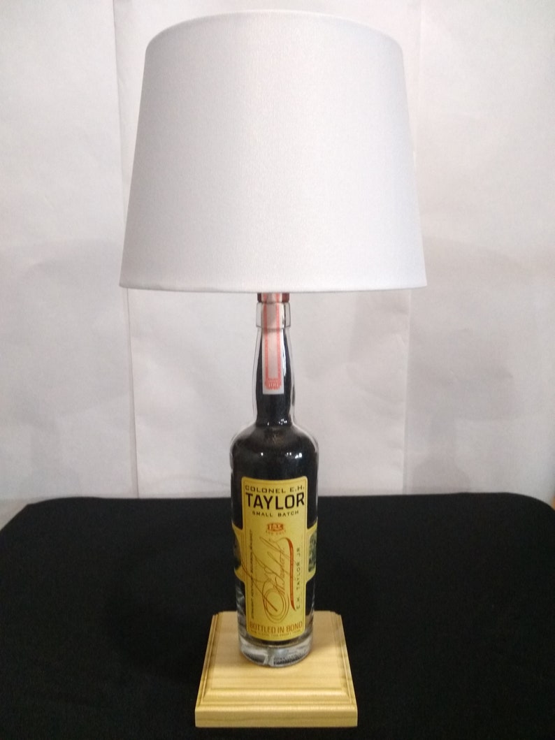 May include: A white lampshade sits atop a clear glass bottle with a black label that reads "Colonel E.H. Taylor Small Batch" and "Bottled in Bond". The bottle is on a wooden base.
