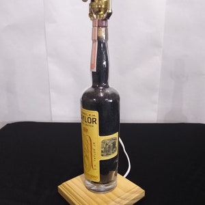 May include: A repurposed bottle lamp with a black and gold label that reads "Colonel E.H. Taylor Jr. Small Batch" and "E.H. Taylor Jr.". The bottle is sitting on a wooden base.