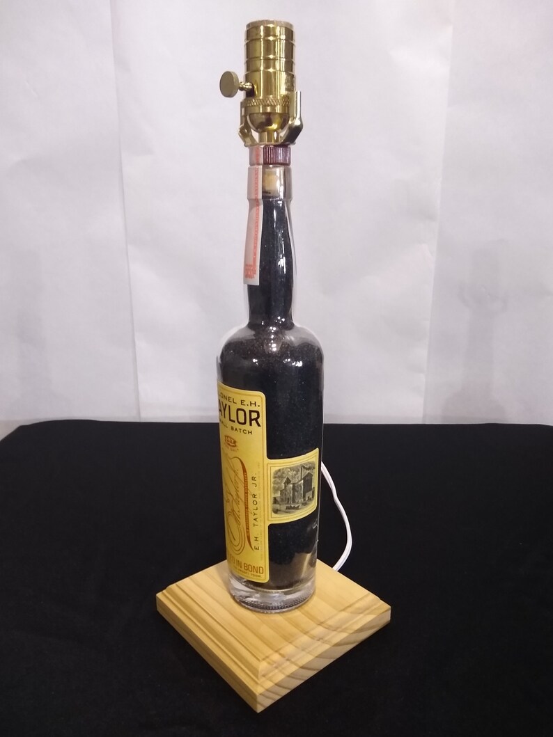 May include: A repurposed bottle lamp with a black and gold label that reads "Colonel E.H. Taylor Jr. Small Batch" and "E.H. Taylor Jr.". The bottle is sitting on a wooden base.