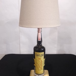 May include: A lamp made from a repurposed bottle of Colonel E.H. Taylor Jr. Small Batch Bourbon Whiskey. The bottle is clear glass with a black label and gold lettering. The lamp has a beige fabric lampshade and a wooden base.