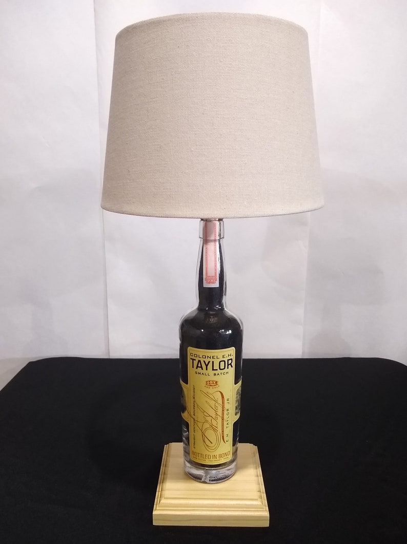 May include: A lamp made from a repurposed bottle of Colonel E.H. Taylor Jr. Small Batch Bourbon Whiskey. The bottle is clear glass with a black label and gold lettering. The lamp has a beige fabric lampshade and a wooden base.