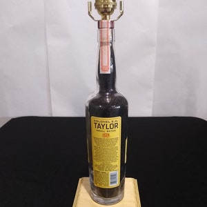 May include: A repurposed bottle lamp made from a Colonel E.H. Taylor Small Batch Bourbon bottle. The bottle is clear glass with a black liquid inside. The lamp has a gold metal lampshade and a wooden base.