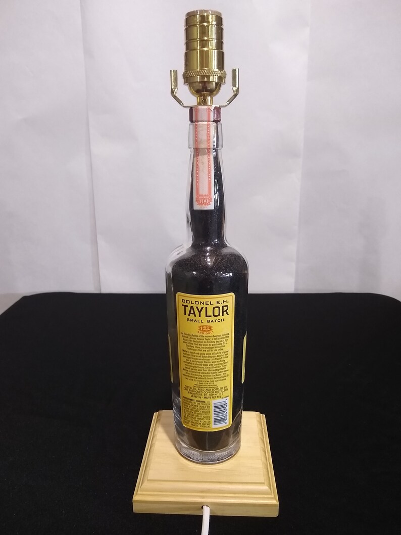 May include: A repurposed bottle lamp made from a Colonel E.H. Taylor Small Batch Bourbon bottle. The bottle is clear glass with a black liquid inside. The lamp has a gold metal lampshade and a wooden base.