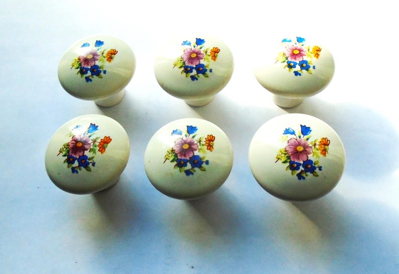 Pu&ograve; includere: Sei pomelli per mobili in ceramica color crema. Ogni pomello presenta un motivo floreale con fiori rosa, blu e arancioni. I pomelli sono rotondi con una base cilindrica corta.