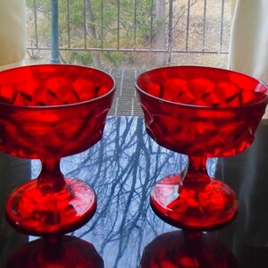 May include: Two ruby red glass dessert bowls with a textured diamond pattern. The bowls have a stemmed base and a wide, shallow bowl. The bowls are displayed on a reflective black surface.