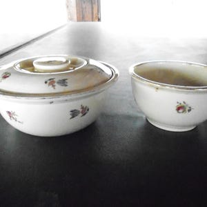 May include: Two white ceramic bowls with floral designs. One bowl has a lid with a knob and is larger than the other. Both bowls have a silver rim and are on a dark surface.