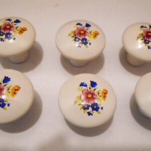 Pu&ograve; includere: Sei pomelli per mobili in ceramica bianco sporco con un motivo floreale. Ogni pomello presenta un fiore rosa centrale, circondato da fiori blu e gialli. I pomelli sono rotondi con una base cilindrica corta.