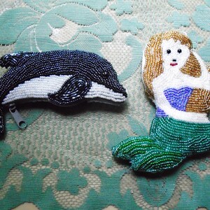 May include: Two beaded coin purses: a black and white dolphin with a zipper, and a mermaid with a gold, white, blue, and green design. The mermaid has gold hair and a blue top.