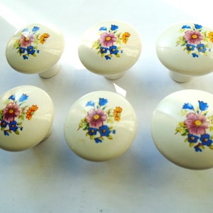 May include: Six off-white ceramic cabinet knobs with floral designs. Each knob features a cluster of purple, blue, and yellow flowers. The knobs are oval-shaped and have a small, cylindrical base.