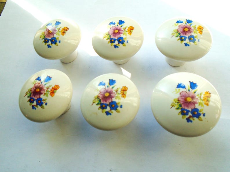 Pu&ograve; includere: Sei pomelli per mobili in ceramica bianco sporco con disegni floreali. Ogni pomello presenta un gruppo di fiori viola, blu e gialli. I pomelli sono di forma ovale e hanno una piccola base cilindrica.