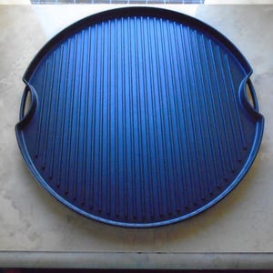 May include: A round, dark blue cast iron grill pan with a ridged cooking surface. The pan has two integrated handles and is designed for stovetop or oven use. The pan is approximately 14 inches in diameter.
