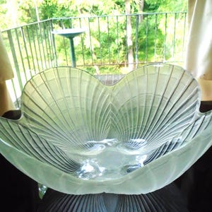 May include: A clear glass bowl featuring a scalloped, flower-like rim and a textured, ribbed pattern radiating from the center base. The bowl has a wide, open design with curved edges that mimic the appearance of a blooming flower petal shape.