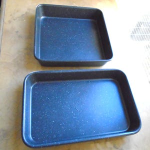 May include: Two rectangular baking pans with a speckled, dark blue finish. The pans have rounded corners and slightly raised edges. The pans are empty and appear to be made of metal.