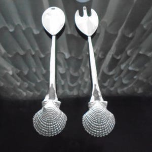 May include: Two silver serving utensils. One is a spoon, the other a fork. Both have long, slender handles and seashell-shaped bases. The utensils are displayed against a dark background.