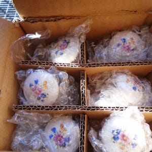 Pu&ograve; includere: Una scatola di cartone contenente sei teiere in ceramica bianca, ciascuna avvolta in plastica trasparente. Le teiere sono decorate con un motivo floreale nei toni del rosa, blu e giallo.