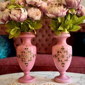 May include: A pair of pink glass vases with a flared rim and pedestal base, each featuring a central gold and green geometric floral pattern. The vases are filled with artificial pink peony flowers and are accompanied by a strand of white pearls.