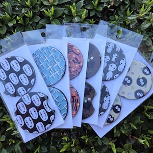May include: Six round, black, cardboard coasters with different colorful designs. The designs include a blue and white cross pattern, a red and white pattern, a black and white star pattern, a black and white pattern with a white creature, and a yellow and black pattern with a black creature.