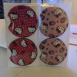 May include: Four car coasters featuring the Hello Kitty character. Two coasters have a red background with white polka dots and Hello Kitty faces. The other two have a pink background with strawberries and Hello Kitty faces.