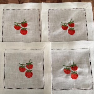 May include: Four white linen coasters with embroidered red tomatoes and green stems. Each coaster has a square shape with a decorative stitched border. The coasters are arranged on a wooden surface.