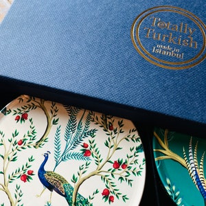 May include: A set of four coasters in a blue box. The coasters feature a peacock design with a white background and green and blue accents. The box has a gold logo that reads "Totally Turkish made in Istanbul."