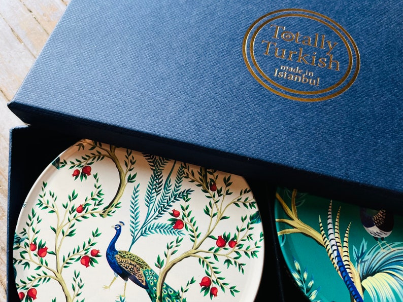 May include: A set of four coasters in a blue box. The coasters feature a peacock design with a white background and green and blue accents. The box has a gold logo that reads "Totally Turkish made in Istanbul."