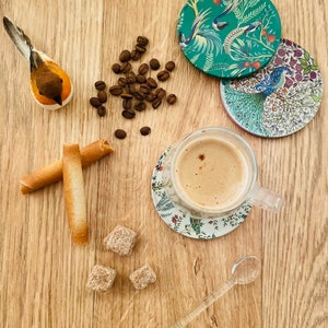 May include: A wooden table with a cup of coffee, two sugar cubes, two breadsticks, a small wooden bird figurine, a pile of coffee beans, and four coasters with floral designs.