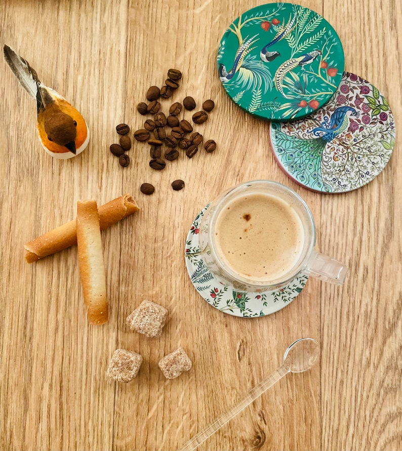 May include: A wooden table with a cup of coffee, two sugar cubes, two breadsticks, a small wooden bird figurine, a pile of coffee beans, and four coasters with floral designs.