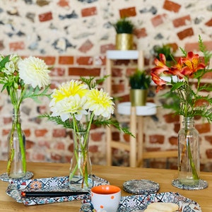 May include: Three ceramic trays with a colorful tile pattern, a white cup with orange polka dots, and two cookies on a wooden table.  The trays are decorated with a variety of floral designs.  There are three vases with flowers in the background.