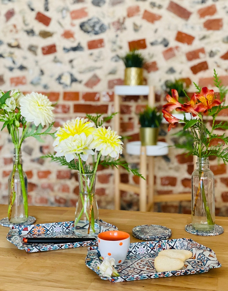 May include: Three ceramic trays with a colorful tile pattern, a white cup with orange polka dots, and two cookies on a wooden table.  The trays are decorated with a variety of floral designs.  There are three vases with flowers in the background.