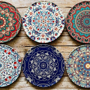 May include: Six round decorative plates with intricate mandala designs in shades of blue, red, green, and white. The plates are arranged in two rows of three on a wooden surface.