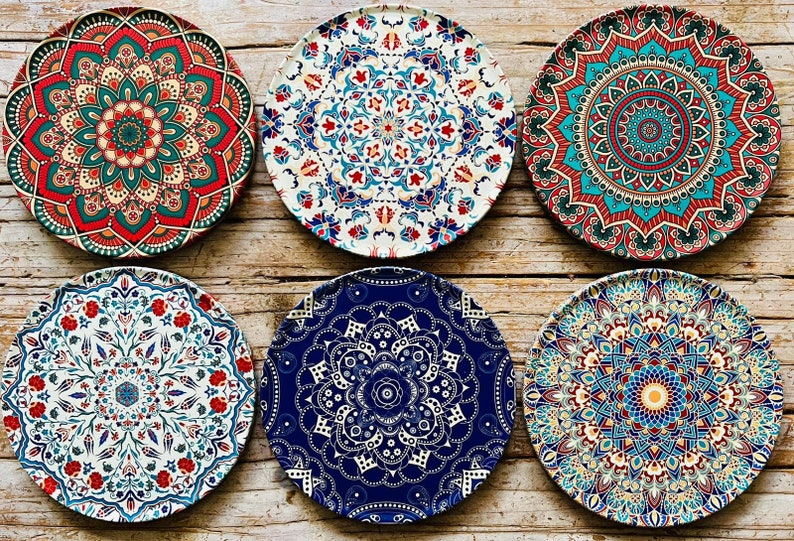 May include: Six round decorative plates with intricate mandala designs in shades of blue, red, green, and white. The plates are arranged in two rows of three on a wooden surface.