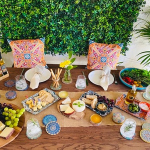 May include: A table setting with a rustic wooden table, a burlap runner, and a variety of decorative elements, including colorful pillows, floral arrangements, and blue and white patterned coasters. The table is set with plates, glasses, and a variety of food items, including cheese, bread, olives, and grapes.