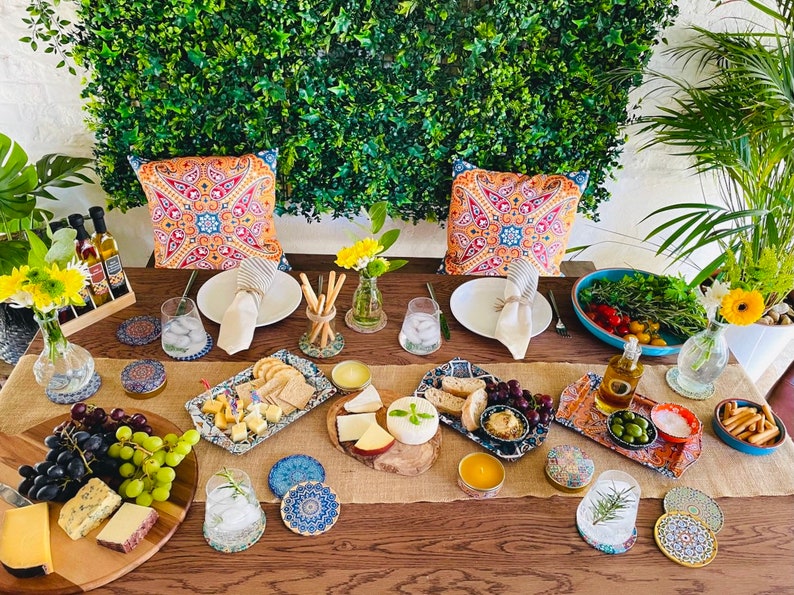 May include: A table setting with a rustic wooden table, a burlap runner, and a variety of decorative elements, including colorful pillows, floral arrangements, and blue and white patterned coasters. The table is set with plates, glasses, and a variety of food items, including cheese, bread, olives, and grapes.