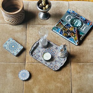 May include: A tray with a blue and white patterned design, featuring a small white candle in the center. The tray is on a beige surface with a wooden bowl, a glass hourglass, a book with the word "Gaudi" on the cover, and a magnifying glass.